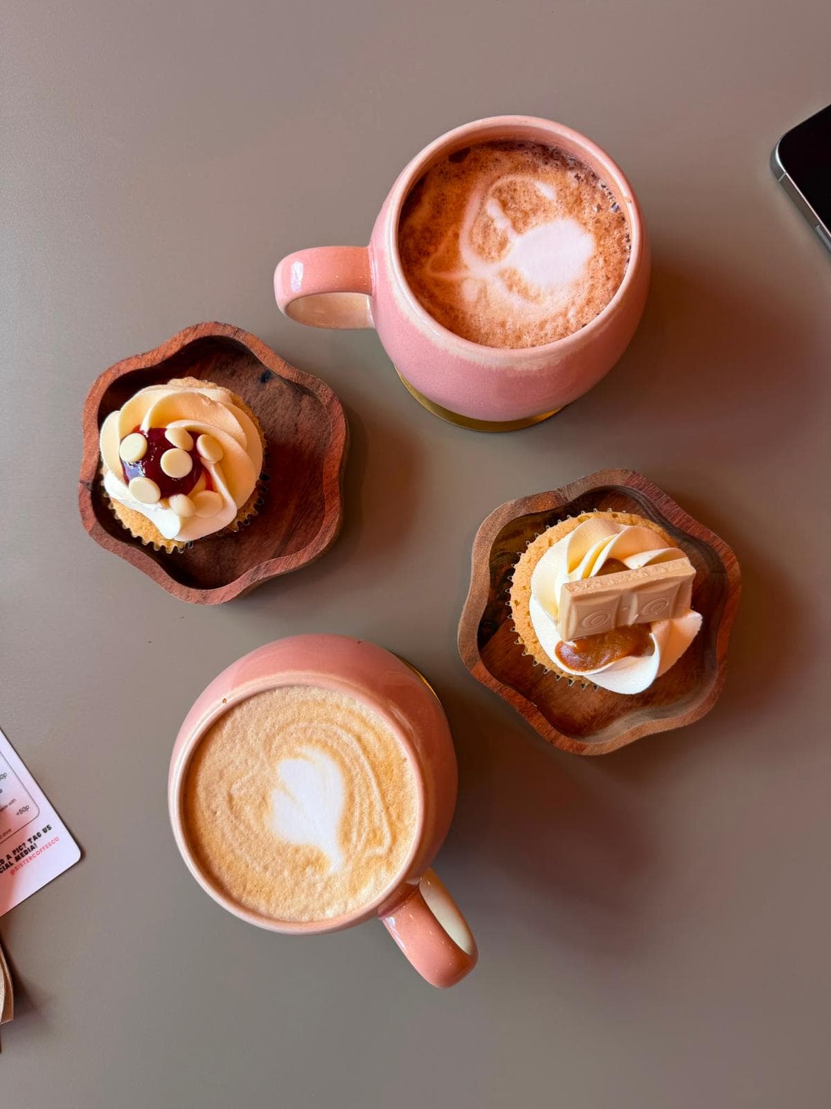 Two pink mugs of coffee beside iced cupcakes on wooden dishes